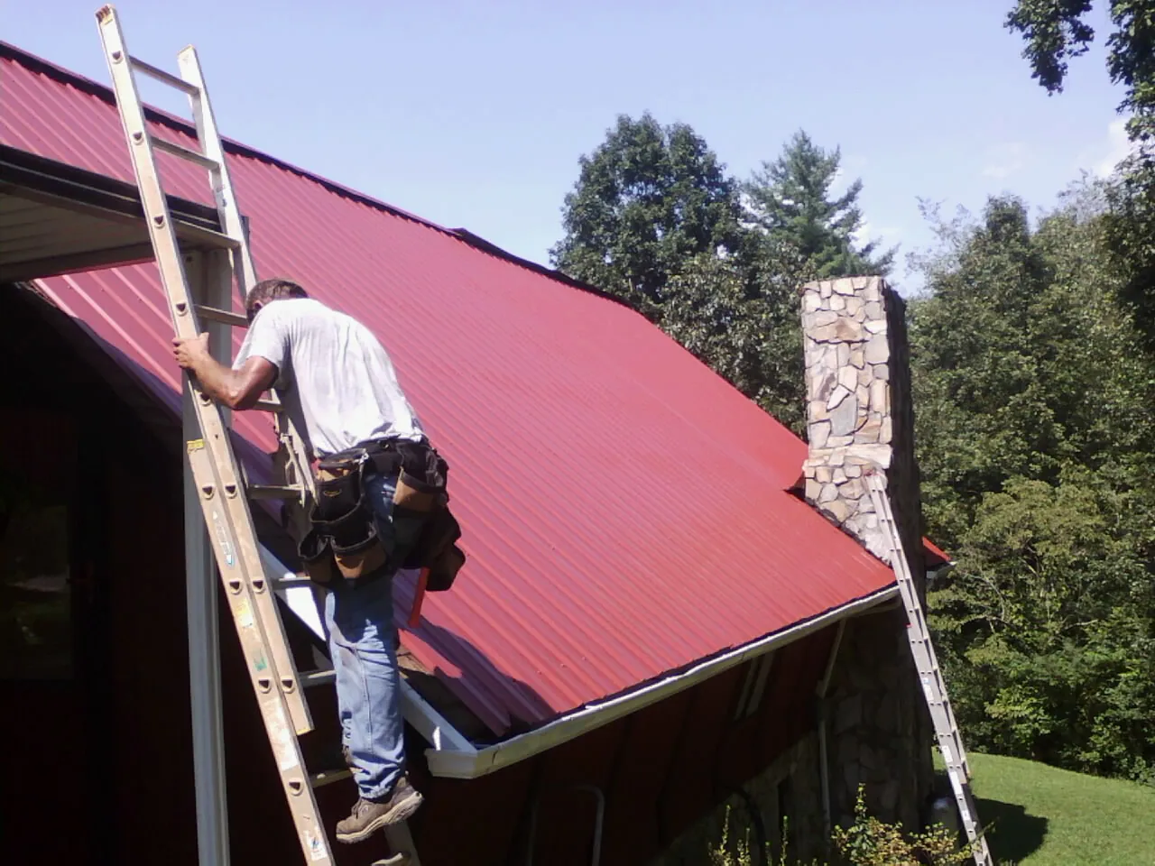 Roofer on a ladder beside a red metal roof and stone chimney — Soffit & Fascia Repair by Roofing Brothers in Kyles Ford